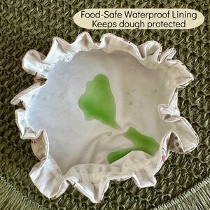 May include: A round fabric bowl cover with a ruffled edge and a white interior lining featuring a green liquid spill. The text Food-Safe Waterproof Lining Keeps dough protected is printed in a beige oval above the bowl cover.