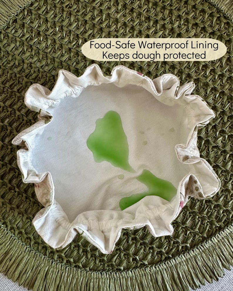 May include: A round fabric bowl cover with a ruffled edge and a white interior lining featuring a green liquid spill. The text Food-Safe Waterproof Lining Keeps dough protected is printed in a beige oval above the bowl cover.