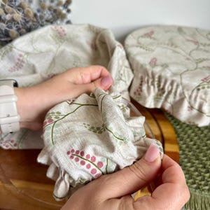 May include: Two reusable fabric bowl covers made from a light beige linen-like material featuring a delicate botanical print of small pink berries and green stems. The covers have an elasticized edge designed to fit snugly over round containers.
