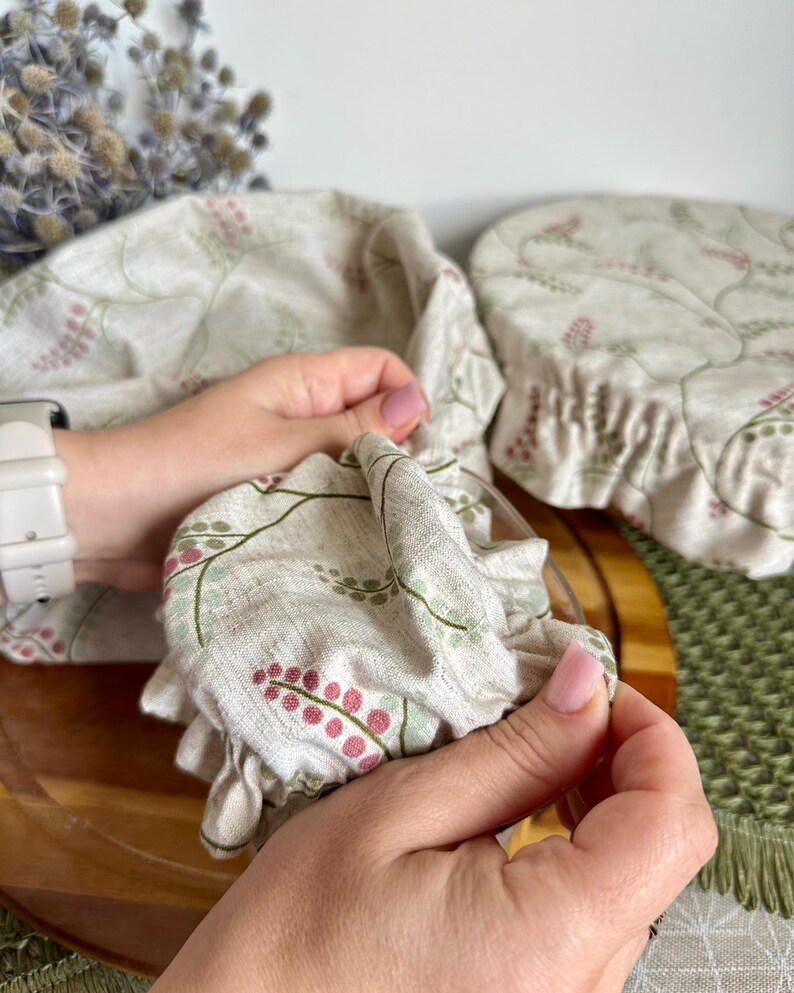 May include: Two reusable fabric bowl covers made from a light beige linen-like material featuring a delicate botanical print of small pink berries and green stems. The covers have an elasticized edge designed to fit snugly over round containers.