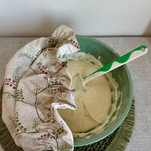 May include: A light green mixing bowl containing thick beige batter with a white and green silicone spatula inside. A beige fabric bowl cover with a botanical print of small red berries and green leaves is draped over one side of the bowl.