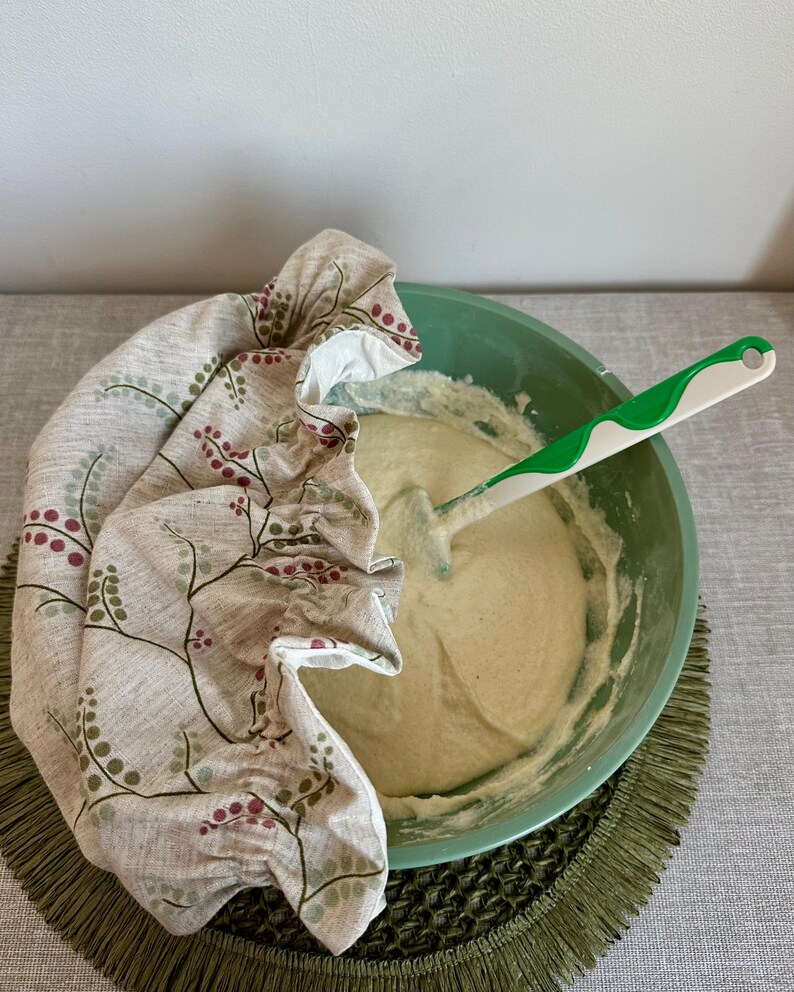 May include: A light green mixing bowl containing thick beige batter with a white and green silicone spatula inside. A beige fabric bowl cover with a botanical print of small red berries and green leaves is draped over one side of the bowl.