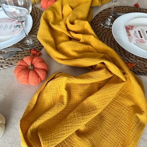 Puede incluir: Un camino de mesa amarillo mostaza sobre una mesa. La mesa está puesta con platos blancos, copas de vino y calabazas decorativas. Los manteles individuales tejidos y un menú se suman al tema otoñal. La tela texturizada del camino de mesa añade interés visual.