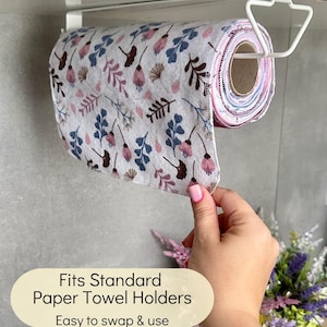 Op de afbeelding: Een rol herbruikbare stoffen doeken met een bloemenpatroon in roze, blauwe en bruine tinten, hangend aan een witte metalen houder onder de kast. De tekst op de afbeelding luidt: Fits Standard Paper Towel Holders, Easy to swap & use.