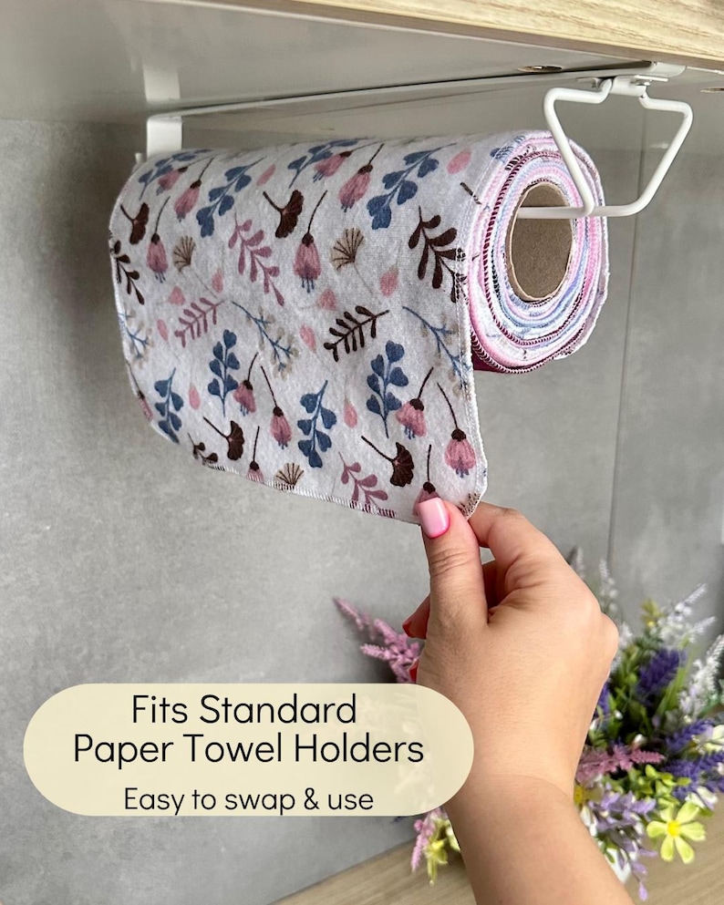 Op de afbeelding: Een rol herbruikbare stoffen doeken met een bloemenpatroon in roze, blauwe en bruine tinten, hangend aan een witte metalen houder onder de kast. De tekst op de afbeelding luidt: Fits Standard Paper Towel Holders, Easy to swap & use.