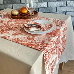 Può includere: Un tavolo da pranzo apparecchiato con un runner in tessuto toile de Jouy rosso e bianco, tovaglioli abbinati e piatti bianchi. Un calice, una piccola zucca e una candela sono su un vassoio di legno. La tovaglia è beige.