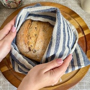 Peut inclure: Un pain rond enveloppé dans une serviette en lin à rayures bleues et blanches. Le pain est posé sur une planche à découper en bois. Le pain a des graines sur le dessus. La serviette est enroulée autour du pain. Le pain est tenu par deux mains.