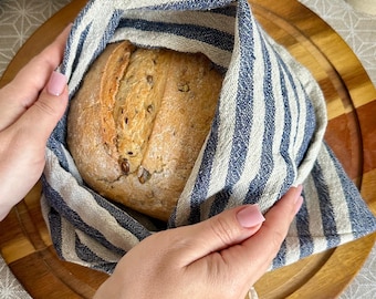 Peut inclure: Une miche de pain artisanal croustillant avec des graines visibles, partiellement enveloppée dans un sac en tissu rayé beige et bleu marine. Le pain présente une croûte dorée avec une entaille distincte sur la surface supérieure.