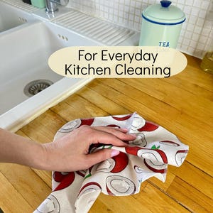 Op de afbeelding: Een vierkante keukendoek met een herhalend patroon van rode en witte kokosnoothelften op een lichte achtergrond. De doek ligt op een houten aanrechtblad. De tekst "For Everyday Kitchen Cleaning" is in het midden van de afbeelding geplaatst.