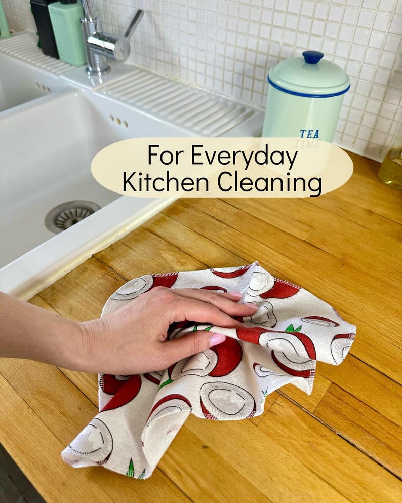 Op de afbeelding: Een vierkante keukendoek met een herhalend patroon van rode en witte kokosnoothelften op een lichte achtergrond. De doek ligt op een houten aanrechtblad. De tekst "For Everyday Kitchen Cleaning" is in het midden van de afbeelding geplaatst.