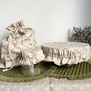 May include: A set of fabric kitchen accessories featuring a beige base with a pattern of small pink berries and green vines. The set includes a drawstring storage bag, a small elasticized bowl cover on a glass jar, and a large elasticized bowl cover.