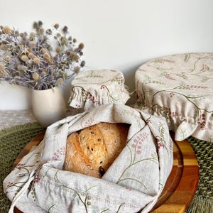 May include: A set of three beige fabric covers and a drawstring bag featuring a delicate botanical pattern of green stems and small pink berries. The drawstring bag holds a round loaf of bread, while the two circular covers sit nearby.