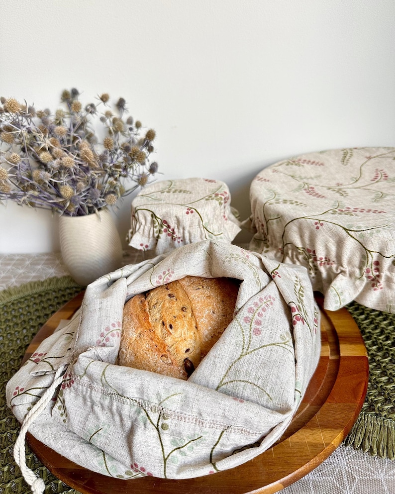 May include: A set of three beige fabric covers and a drawstring bag featuring a delicate botanical pattern of green stems and small pink berries. The drawstring bag holds a round loaf of bread, while the two circular covers sit nearby.
