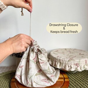 May include: A beige fabric bread bag with a drawstring closure and a botanical pattern of small red berries and green stems. The text on the image reads Drawstring Closure & Keeps bread fresh. A matching fabric bowl cover is also visible.