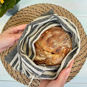 May include: A round, crusty loaf of artisan bread with a golden-brown, flour-dusted surface, nestled inside a reusable fabric bag featuring a black and beige vertical striped pattern with a textured, woven appearance and a drawstring closure.