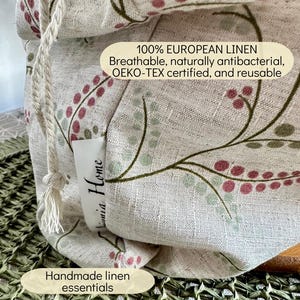 May include: A beige linen drawstring bag featuring a botanical pattern of green stems and pink berries. A white fabric tag reads "Linnia Home". Text on the bag states: "100% EUROPEAN LINEN Breathable, naturally antibacterial, OEKO-TEX certified, and reusable".