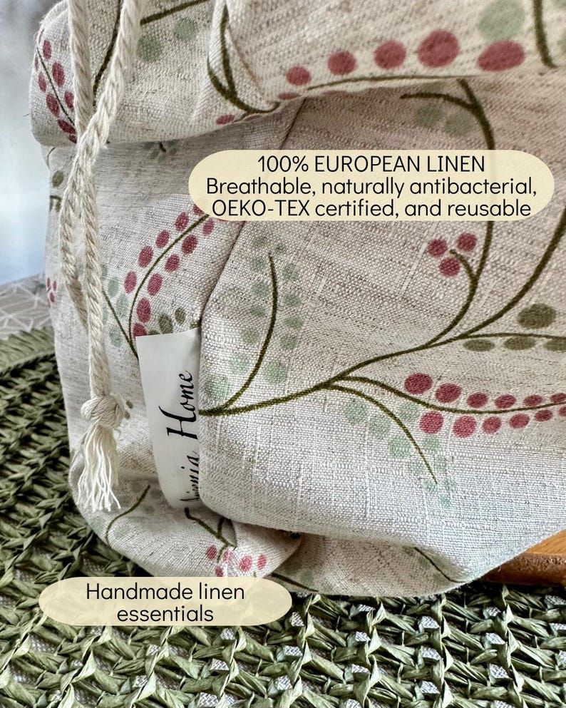 May include: A beige linen drawstring bag featuring a botanical pattern of green stems and pink berries. A white fabric tag reads "Linnia Home". Text on the bag states: "100% EUROPEAN LINEN Breathable, naturally antibacterial, OEKO-TEX certified, and reusable".