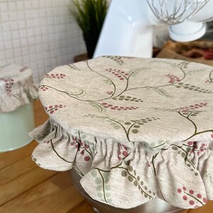 May include: A round fabric bowl cover with an elasticized ruffled edge, featuring a beige base with a repeating pattern of thin dark green stems and small red and green berries. The cover is fitted over a stainless steel mixing bowl.