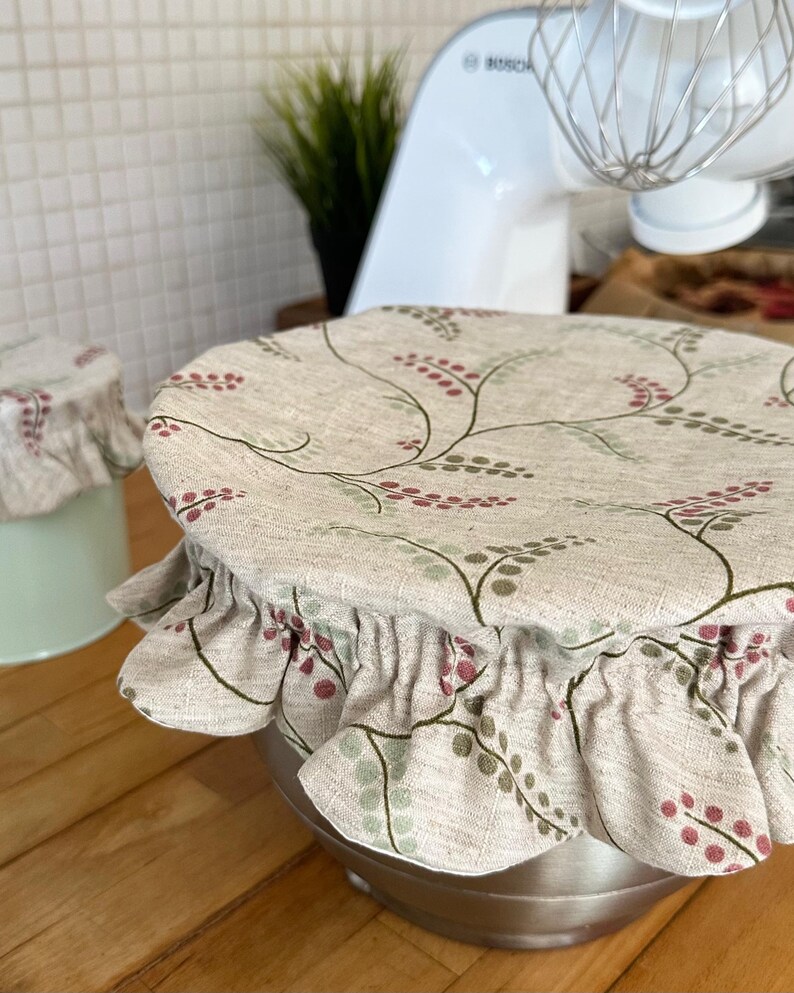 May include: A round fabric bowl cover with an elasticized ruffled edge, featuring a beige base with a repeating pattern of thin dark green stems and small red and green berries. The cover is fitted over a stainless steel mixing bowl.