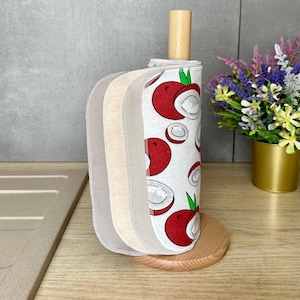 Op de afbeelding: Een set herbruikbare stoffen doekjes gestapeld op een lichthouten keukenrolhouder. De doekjes hebben een witte achtergrond met een herhalend patroon van rode en witte lychees met groene blaadjes, naast effen beige en lichtgrijze stoffen doekjes.