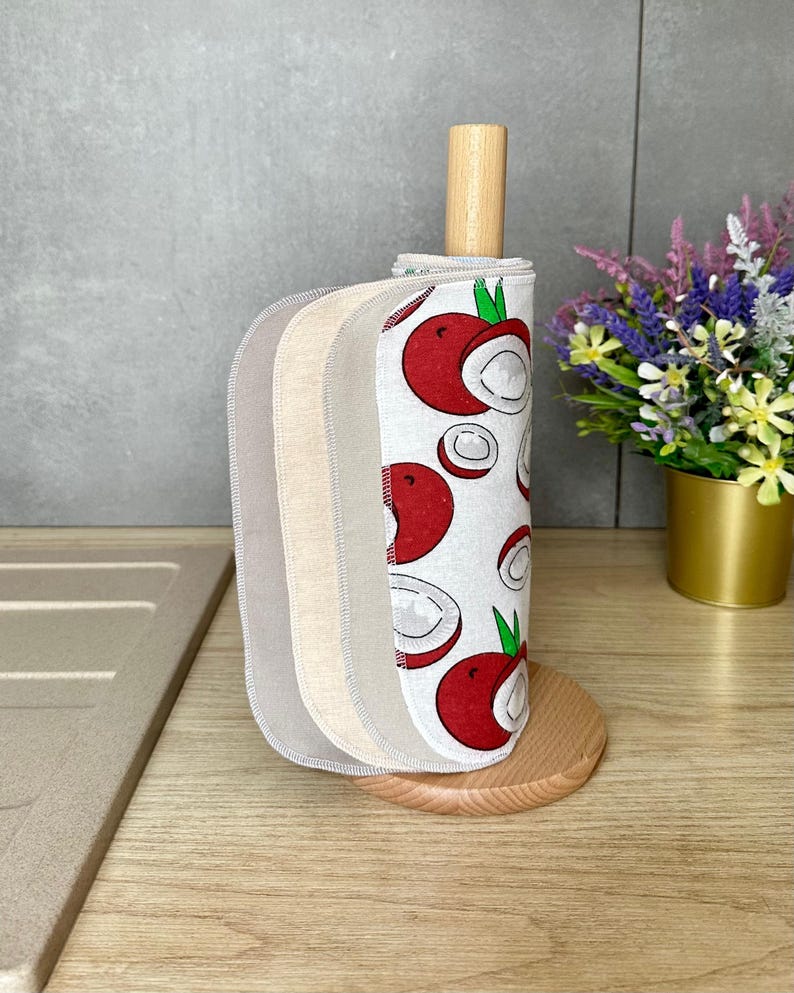Op de afbeelding: Een set herbruikbare stoffen doekjes gestapeld op een lichthouten keukenrolhouder. De doekjes hebben een witte achtergrond met een herhalend patroon van rode en witte lychees met groene blaadjes, naast effen beige en lichtgrijze stoffen doekjes.