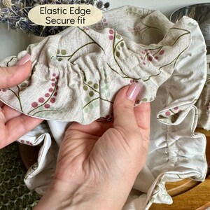 May include: A beige fabric bowl cover featuring a botanical print of small pink and green berries on thin stems. The cover has an elastic edge for a secure fit, as indicated by the text Elastic Edge Secure fit printed on the image.