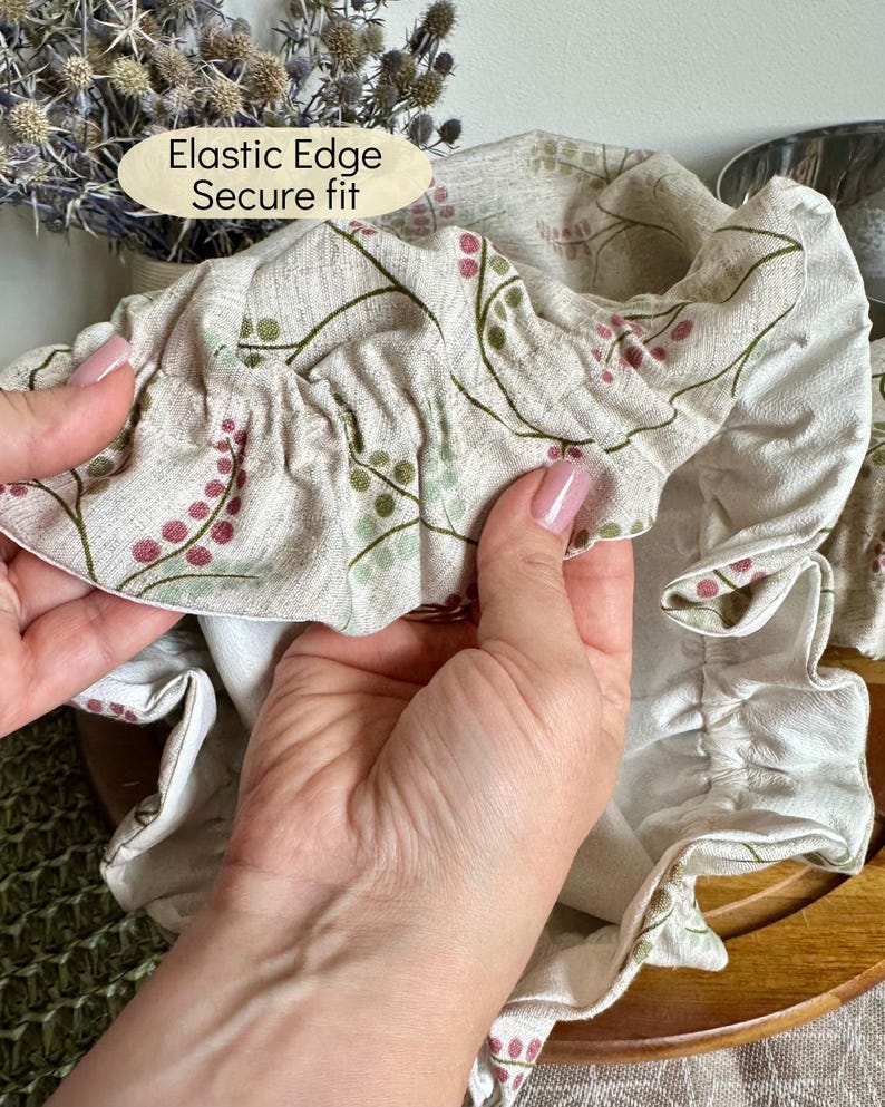 May include: A beige fabric bowl cover featuring a botanical print of small pink and green berries on thin stems. The cover has an elastic edge for a secure fit, as indicated by the text Elastic Edge Secure fit printed on the image.