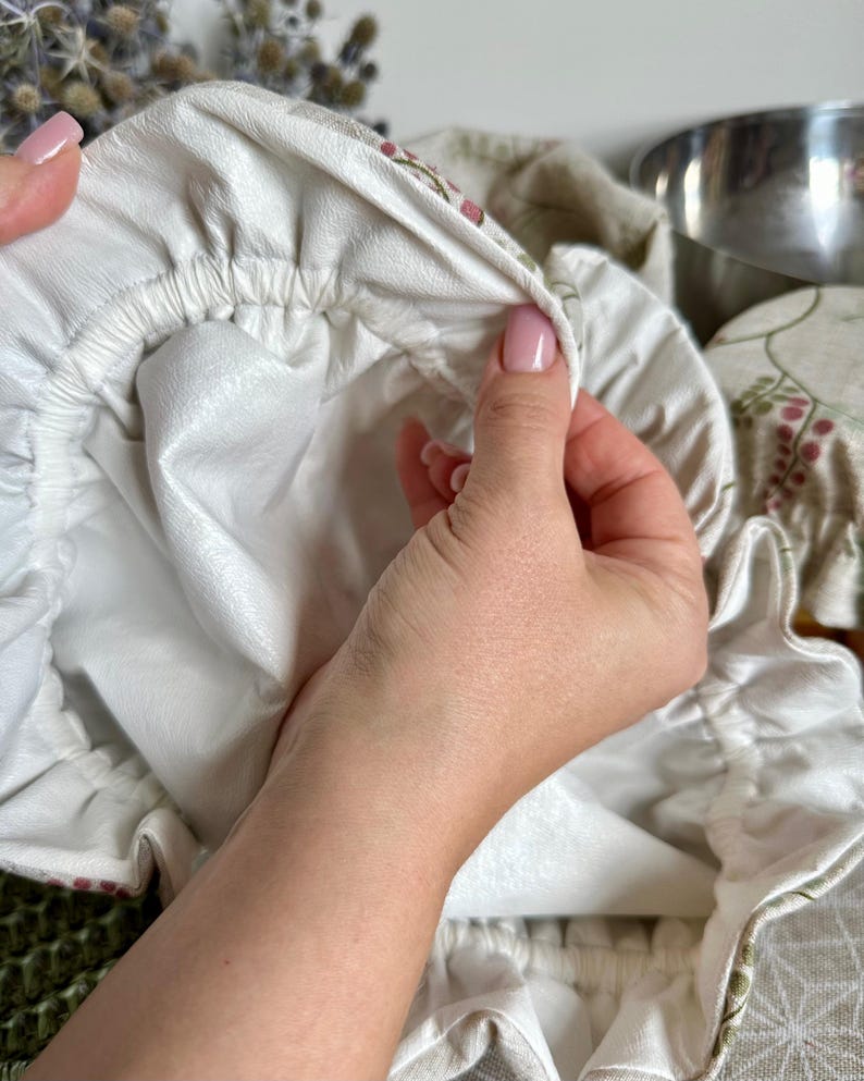 May include: A round fabric bowl cover featuring a beige exterior with a delicate floral pattern of small pink berries and green leaves. The interior is lined with a smooth, solid white material, and the edge is finished with an elastic band.