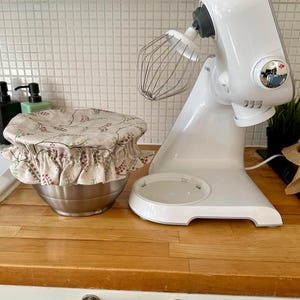 May include: A white stand mixer with a metal whisk attachment positioned next to a stainless steel mixing bowl covered by a beige fabric bowl cover featuring a repeating pattern of small red and green leaves and stems with a ruffled elastic edge.