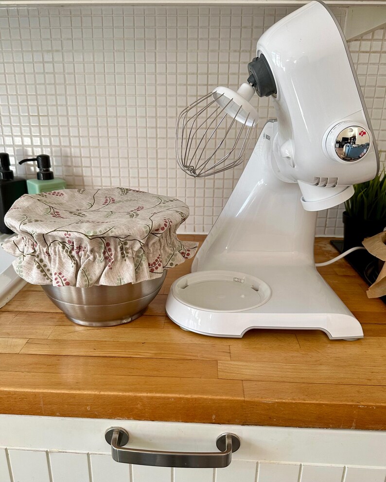 May include: A white stand mixer with a metal whisk attachment positioned next to a stainless steel mixing bowl covered by a beige fabric bowl cover featuring a repeating pattern of small red and green leaves and stems with a ruffled elastic edge.
