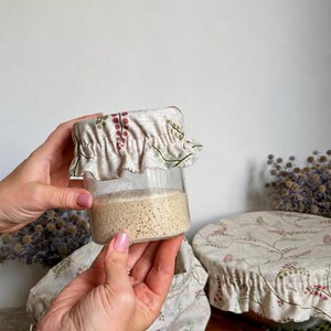 May include: A clear glass jar filled with beige sourdough starter, topped with a beige fabric cover featuring a delicate green and pink botanical print. The fabric cover has an elasticized, ruffled edge that fits snugly around the jar rim.