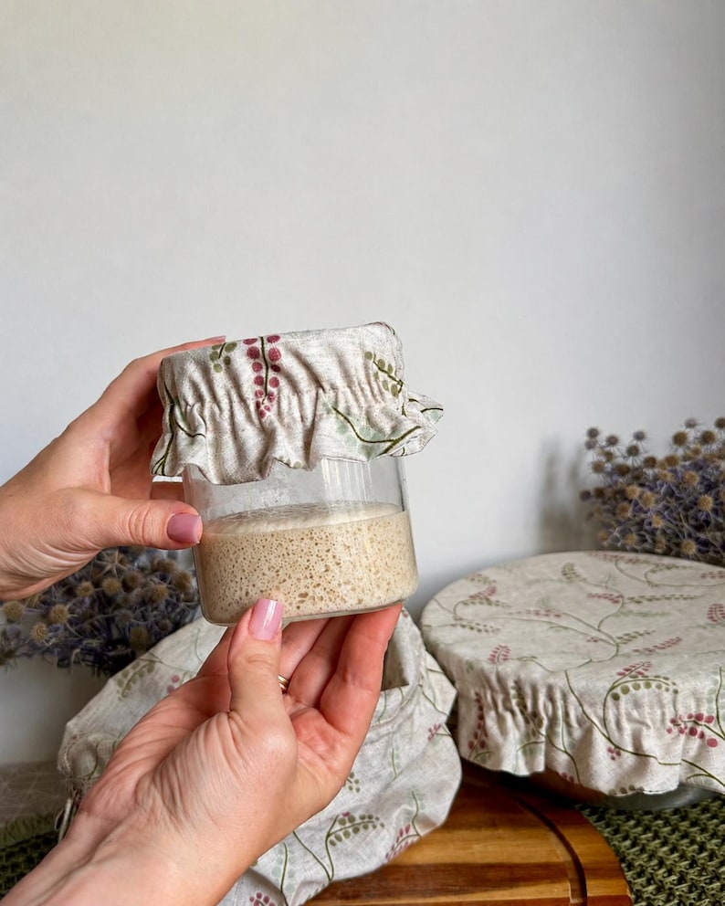 May include: A clear glass jar filled with beige sourdough starter, topped with a beige fabric cover featuring a delicate green and pink botanical print. The fabric cover has an elasticized, ruffled edge that fits snugly around the jar rim.