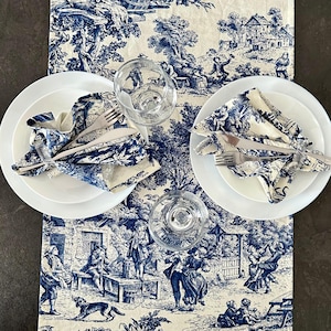 May include: Two white plates with blue and white patterned napkins folded on top. The plates are set on a white tablecloth with a blue and white toile pattern. The tablecloth features a scene of people in a garden setting.