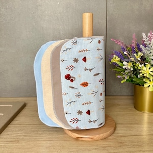 Op de afbeelding: Een lichtblauwe houten keukenrolhouder met een stapel herbruikbare stoffen doeken. De doeken hebben een patroon van kleine rode paddenstoelen, bruine bladeren en takjes. De stapel bevat effen lichtblauwe, beige en bruine stoffen vellen.