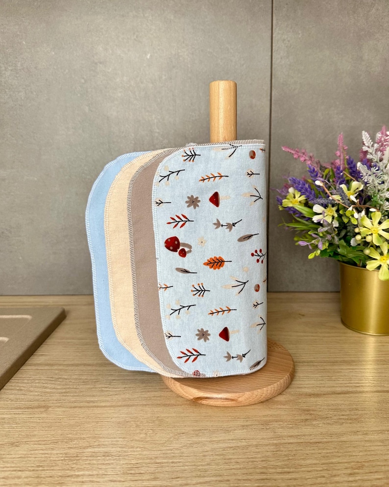 Op de afbeelding: Een lichtblauwe houten keukenrolhouder met een stapel herbruikbare stoffen doeken. De doeken hebben een patroon van kleine rode paddenstoelen, bruine bladeren en takjes. De stapel bevat effen lichtblauwe, beige en bruine stoffen vellen.