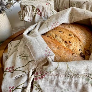 May include: A beige linen bread bag and matching bowl covers featuring a botanical print of small pink berries and green leaves. The bread bag is open, revealing a loaf of crusty, seeded bread inside, while the covers are gathered with elastic.