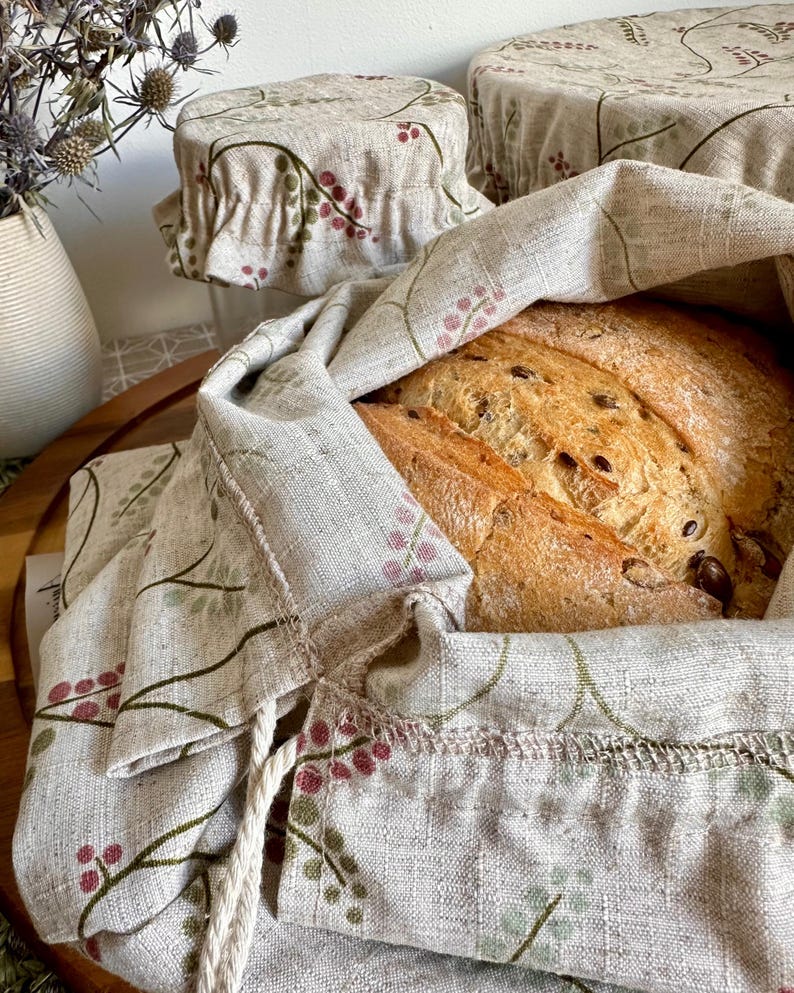 May include: A beige linen bread bag and matching bowl covers featuring a botanical print of small pink berries and green leaves. The bread bag is open, revealing a loaf of crusty, seeded bread inside, while the covers are gathered with elastic.