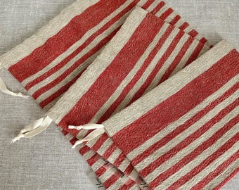 Puede incluir: Cuatro bolsas de lino a rayas rojas y beige dispuestas en diagonal sobre una superficie gris claro. Cada bolsa tiene una cinta blanca. Las bolsas tienen una estética rústica y campestre.