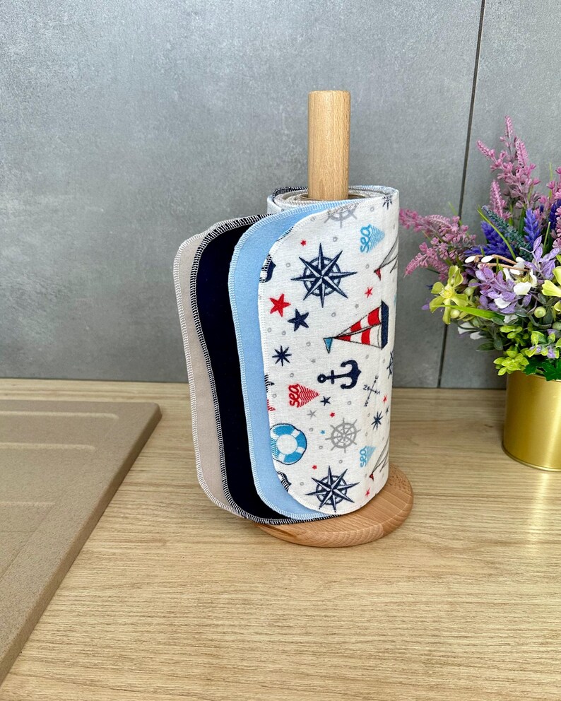 Op de afbeelding: Een houten keukenrolhouder met een rol herbruikbare stoffen doekjes. De buitenste doek heeft een nautisch patroon met zeilboten, ankers, kompassen en reddingsboeien in rood, blauw en grijs op een witte ondergrond.