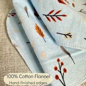 Op de afbeelding: Een lichtblauwe katoenen flanellen doek met een herhalend patroon van kleine bruine, oranje en cr&egrave;mekleurige bladeren, bessen en bloemen. De stof heeft met de hand afgewerkte randen met een lichtblauwe locksteek en de tekst 100% Cotton Flannel Hand-finished edges.