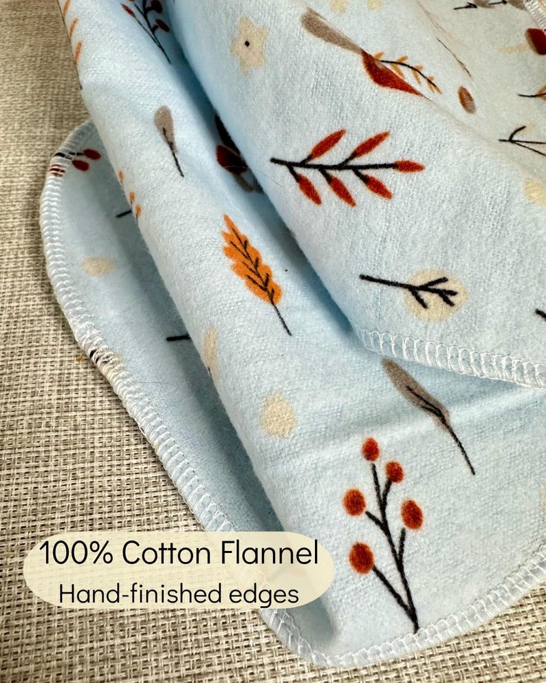 Op de afbeelding: Een lichtblauwe katoenen flanellen doek met een herhalend patroon van kleine bruine, oranje en cr&egrave;mekleurige bladeren, bessen en bloemen. De stof heeft met de hand afgewerkte randen met een lichtblauwe locksteek en de tekst 100% Cotton Flannel Hand-finished edges.