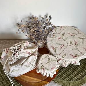 May include: A set of three fabric items featuring a beige background with a repeating pattern of green stems and small pink berries. The set includes a drawstring bag, a small circular bowl cover, and a large circular bowl cover with elastic.