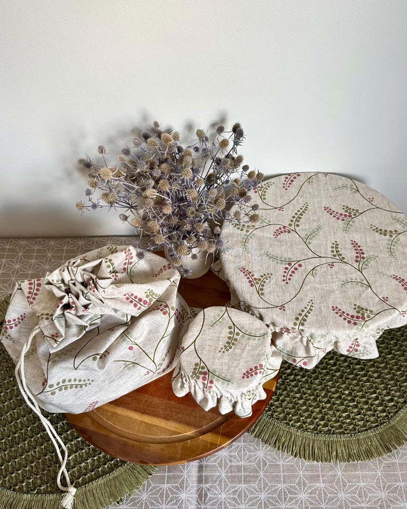 May include: A set of three fabric items featuring a beige background with a repeating pattern of green stems and small pink berries. The set includes a drawstring bag, a small circular bowl cover, and a large circular bowl cover with elastic.