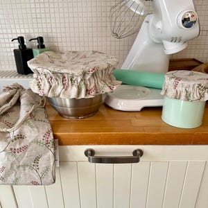 May include: A set of fabric bowl covers featuring a beige background with a repeating pattern of small green leaves and pink berries. The set includes two elasticized bowl covers and a matching drawstring bag, all made from the same floral print fabric.