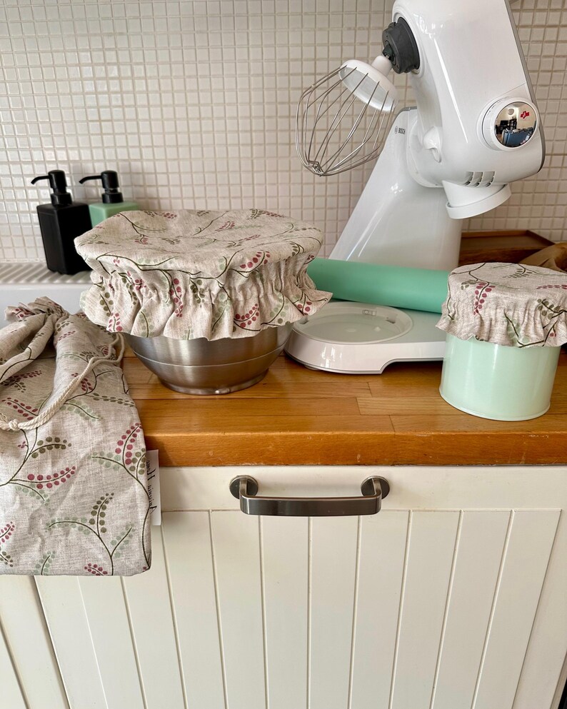 May include: A set of fabric bowl covers featuring a beige background with a repeating pattern of small green leaves and pink berries. The set includes two elasticized bowl covers and a matching drawstring bag, all made from the same floral print fabric.