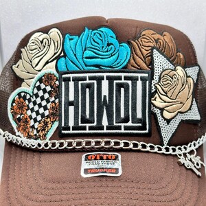 May include: Brown trucker hat with a white "HOWDY" patch, a blue rose, a white rose, a brown rose, a checkered heart, and a silver star with a white rose. The hat has a silver chain with a charm attached.