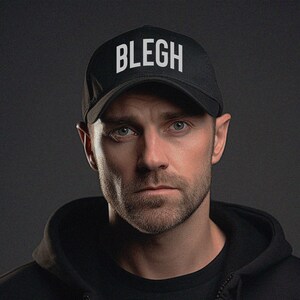 Könnte beinhalten: Ein Mann trägt einen schwarzen Hoodie und eine schwarze Baseballkappe mit dem Wort "BLEGH" in weißen Buchstaben auf der Vorderseite.