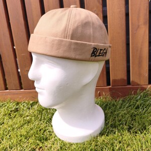Könnte beinhalten: Eine beige, randlose Mütze mit dem Wort "BLEGH" in Schwarz auf der Vorderseite gestickt. Die Mütze wird auf einem weißen Schaufensterkopf vor einem Holzzaun und grünem Gras präsentiert.