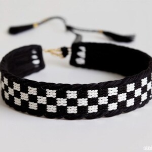 May include: Black and white checkered pattern bracelet. The bracelet is made of woven material and features a repeating pattern of black and white squares. The bracelet has a sliding knot closure.