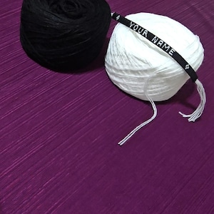 May include: Two yarn balls, one black and one white, sit on a purple surface. A black band with the text "YOUR NAME" in white is wrapped around the white yarn. The yarn is likely for crafting projects.